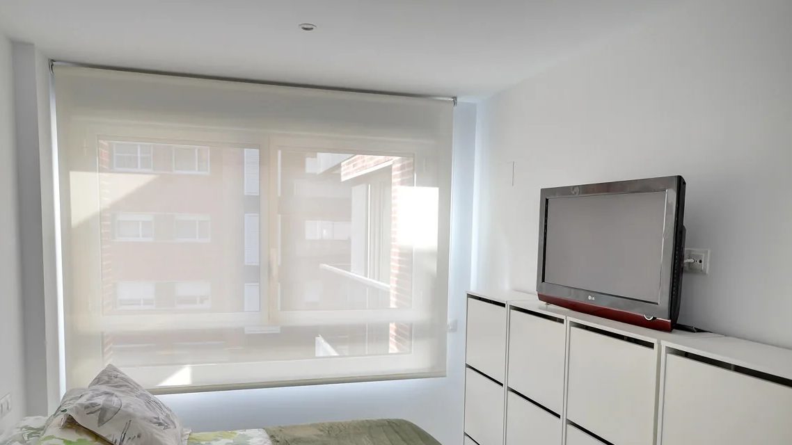 Dormitorio con un estor enrollable screen Javea 5 blanco que deja ver los edificios de enfrente