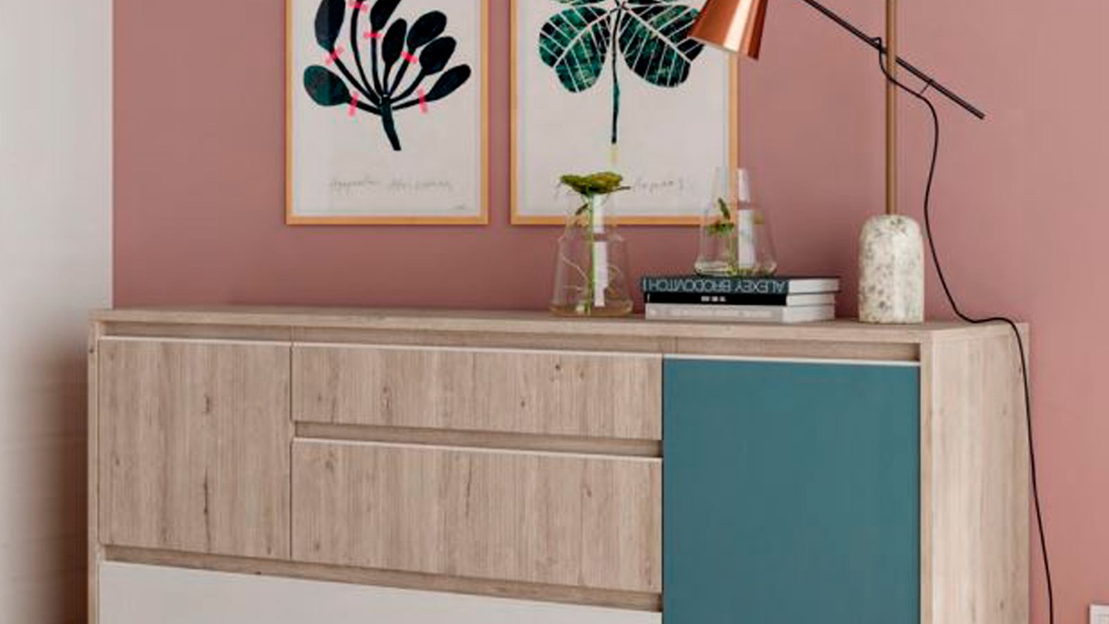 Aparador de madera clara con una puerta azul petróleo en un salón de pared rosa, con dos láminas y un flexo encima